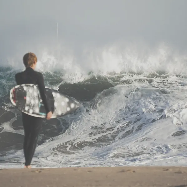 Surfer with a board watching a breaking wave, shown flat with low contrast and muted colour