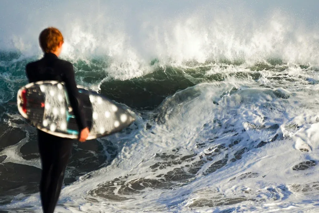 Surfer with a board under the arm watching waves break on the shore