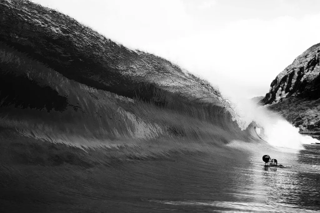 Black and white photo of a photographer in the water under a large breaking wave