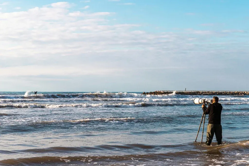 Surf photographer with a long telephoto lens on a tripod, standing in the shallows