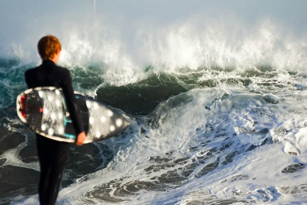 Surfer in a wetsuit holding a board, watching a heavy wave break