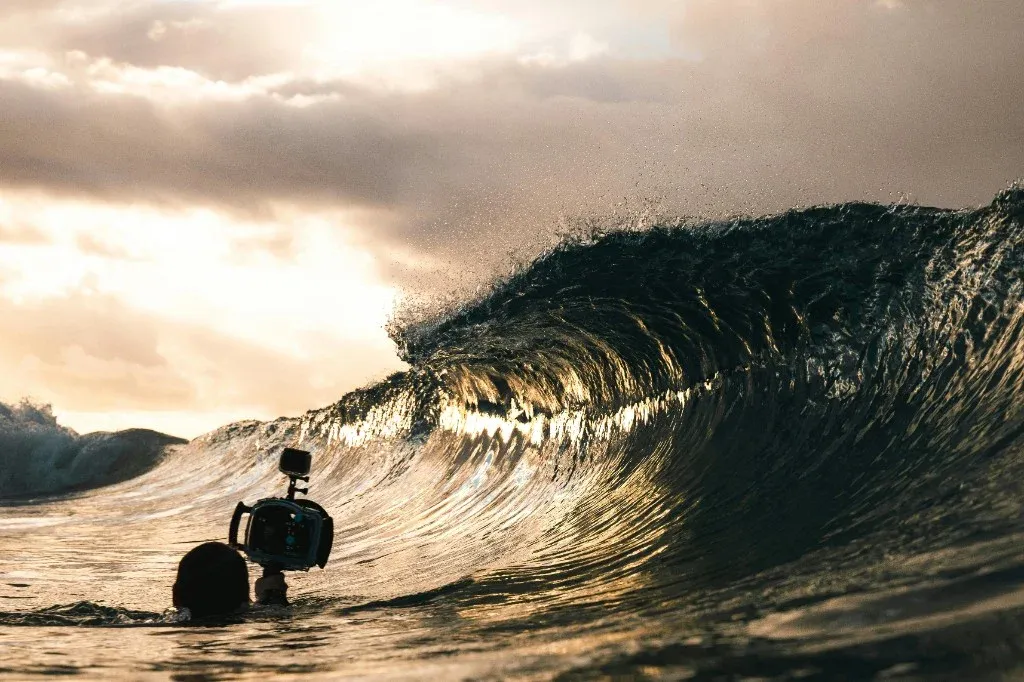 Camera in a water housing at the waterline, backlit by a low sun over a wave